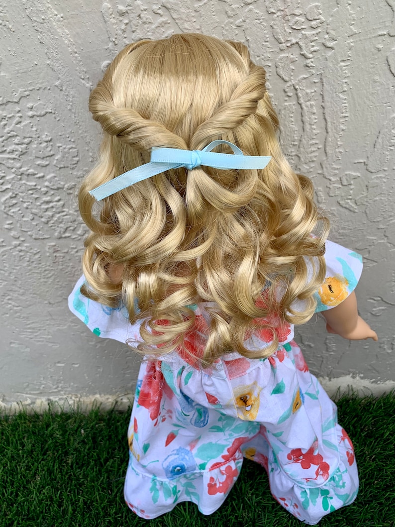 May include: A doll with blonde, curly hair styled with a twisted crown and a light blue ribbon. The doll is wearing a white floral print jumpsuit with red, yellow, and blue flowers.