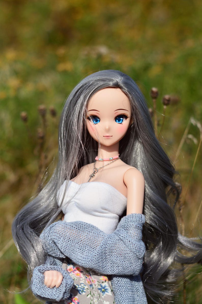 May include: A doll with long, silver hair, blue eyes, and a light pink blush. She is wearing a white tank top, a light blue knitted cardigan, and a floral skirt.