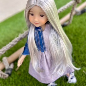 May include: A doll with long, silver hair and brown eyes, wearing a denim jacket over a lavender dress and white shoes. The doll is positioned on a green surface with a rope in the background.