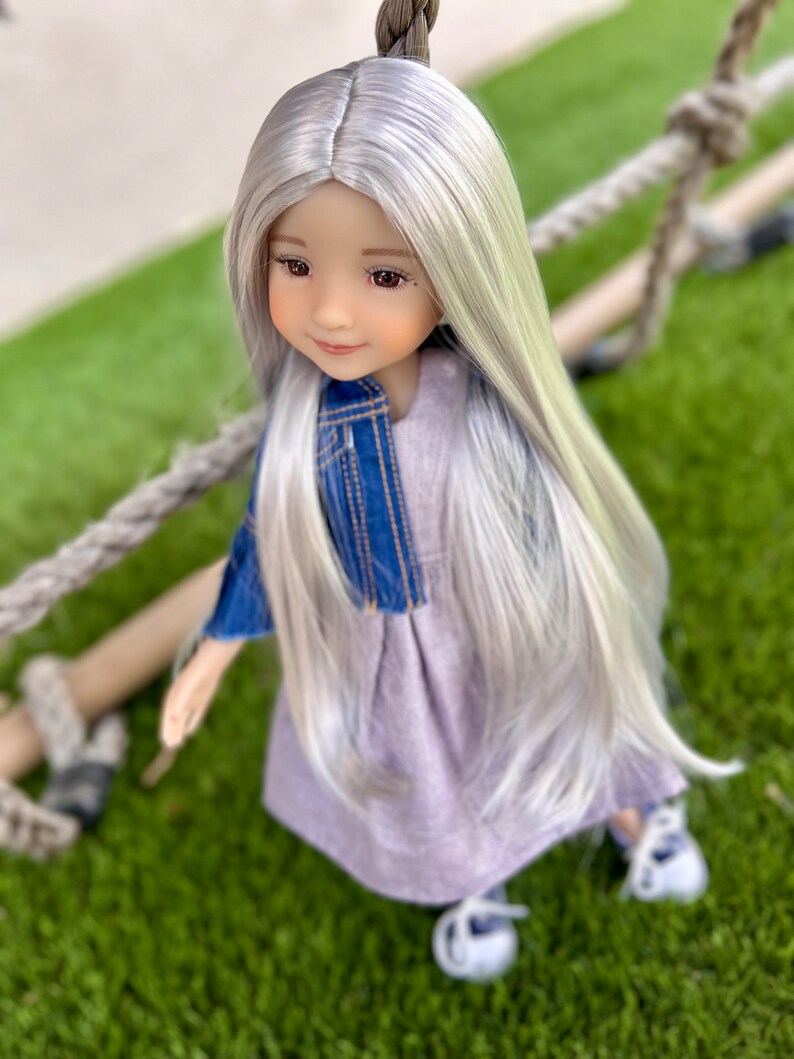 May include: A doll with long, silver hair and brown eyes, wearing a denim jacket over a lavender dress and white shoes. The doll is positioned on a green surface with a rope in the background.