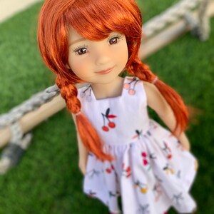 May include: A red-haired doll wearing a white dress with a cherry print. The doll has brown eyes and a sweet smile.