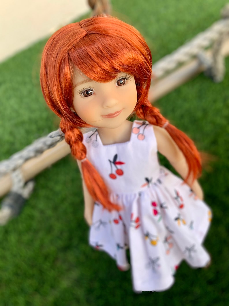May include: A red-haired doll wearing a white dress with a cherry print. The doll has brown eyes and a sweet smile.