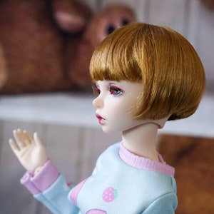 May include: A doll with short, straight, light brown hair and blue eyes. The doll is wearing a light blue and pink sweater with a strawberry design.