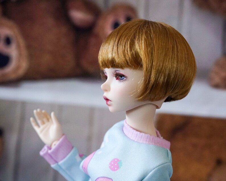 May include: A doll with short, straight, light brown hair and blue eyes. The doll is wearing a light blue and pink sweater with a strawberry design.