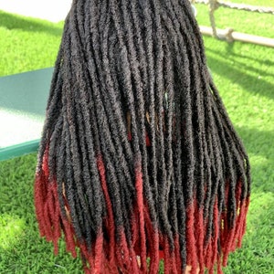 May include: A head of black dreadlocks with red tips. The dreadlocks are long and hang down the back. The hair is styled with a natural look, and the red tips add a pop of colour. The person is wearing checkered shoes.