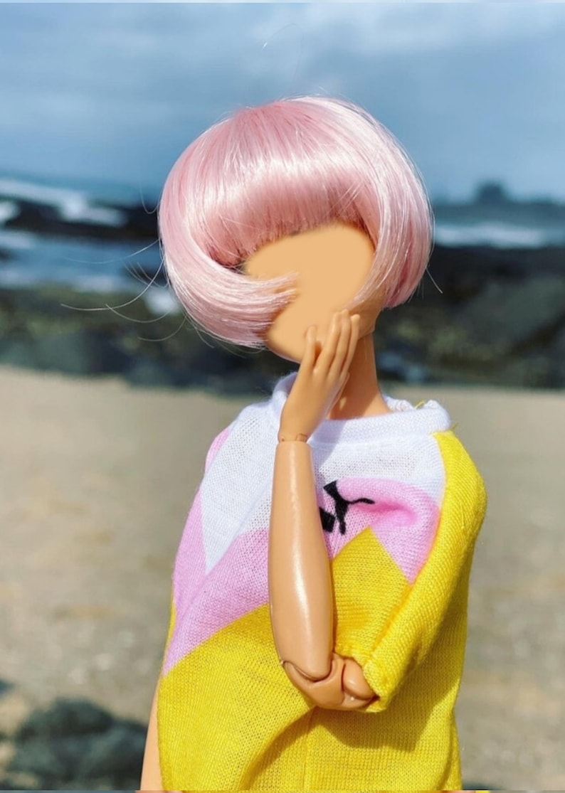 Peut inclure: Une poup&eacute;e aux cheveux roses portant un pull ray&eacute; jaune, rose et blanc. Le visage de la poup&eacute;e est cach&eacute; par sa main.