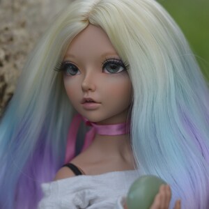 May include: A doll with long, wavy, multi-colored hair, wearing a white shirt and a pink ribbon around her neck. She is holding a green stone in her hand.