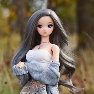 May include: A doll with long, gray hair, wearing a white top, a gray cardigan, and a floral skirt. The doll has blue eyes and is standing in a grassy area with a blurred background of trees.
