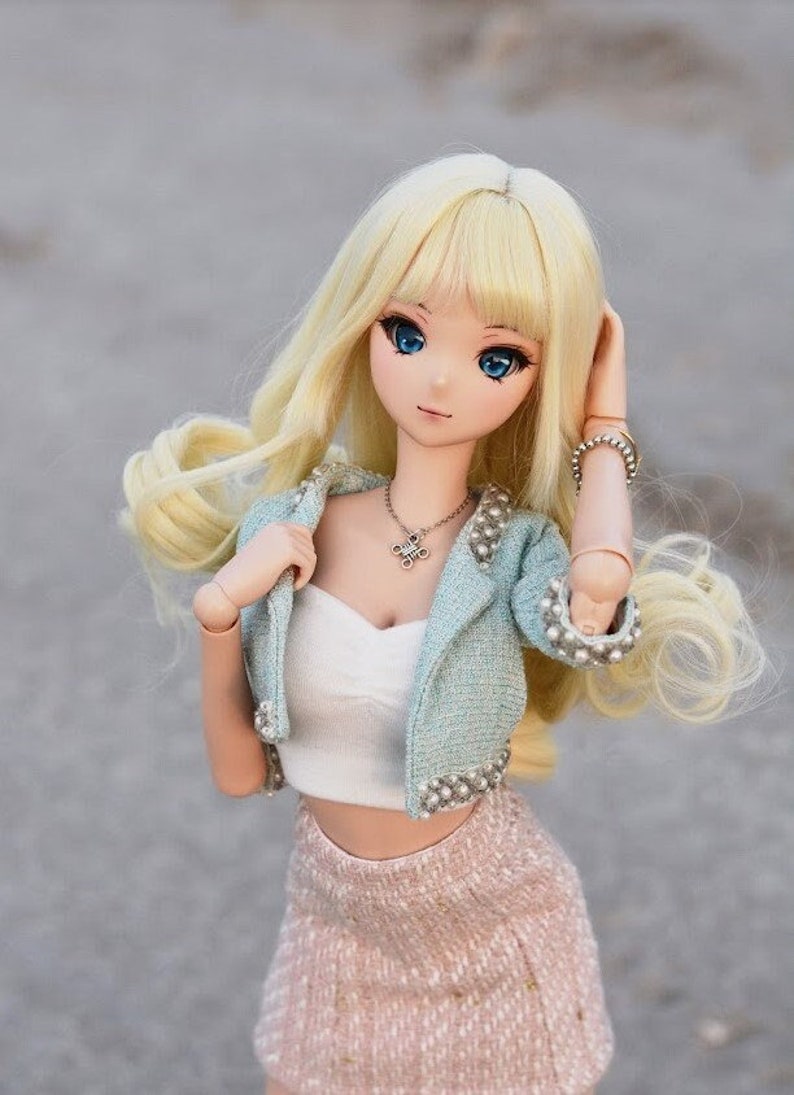May include: A blonde doll with blue eyes wearing a light blue jacket with pearl accents, a white crop top, and a pink skirt.