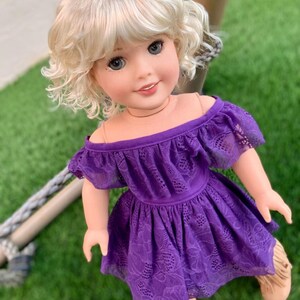 May include: A doll with blonde curly hair wearing a purple off-the-shoulder dress with lace detailing.
