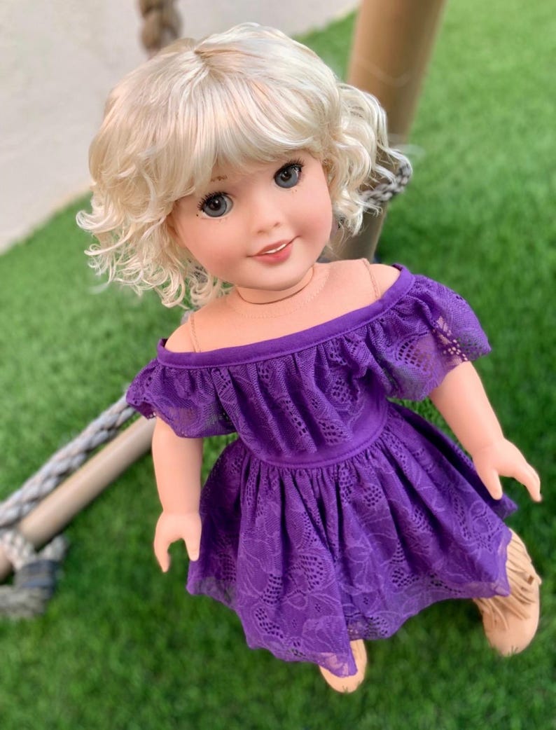 May include: A doll with blonde curly hair wearing a purple off-the-shoulder dress with lace detailing.