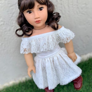 May include: A white lace off-the-shoulder top and skirt set for a doll. The doll has brown hair and brown eyes. The outfit is perfect for a special occasion.
