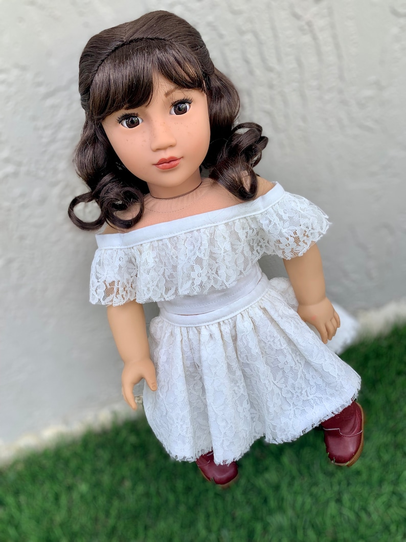 May include: A white lace off-the-shoulder top and skirt set for a doll. The doll has brown hair and brown eyes. The outfit is perfect for a special occasion.