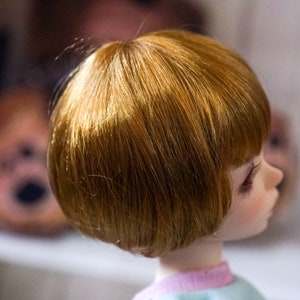 May include: A doll with short, straight, light brown hair. The doll is wearing a light blue shirt with a pink stripe.