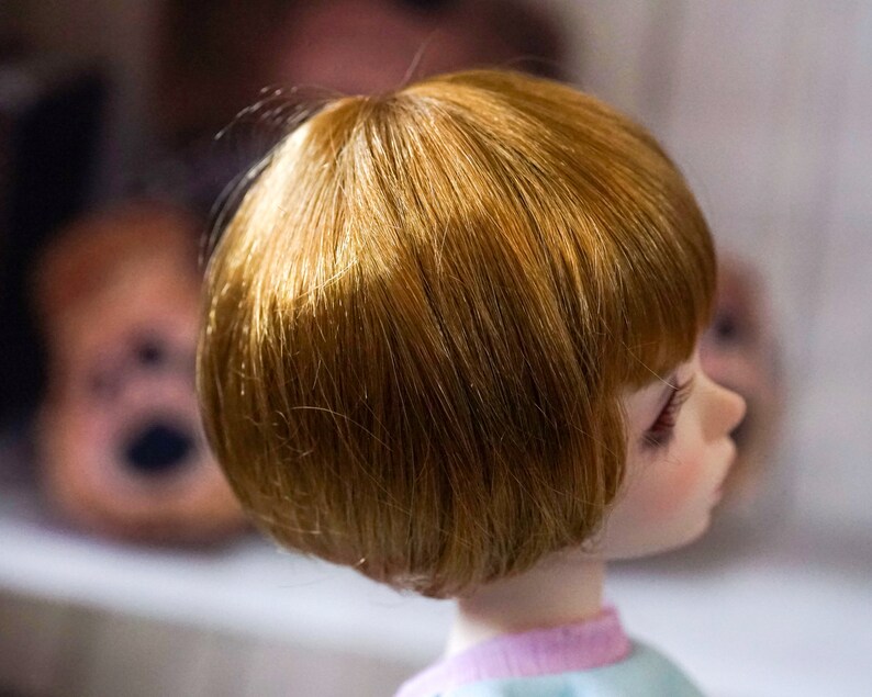May include: A doll with short, straight, light brown hair. The doll is wearing a light blue shirt with a pink stripe.
