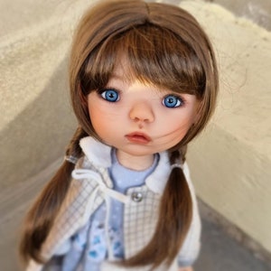 Puede incluir: Una muñeca de porcelana con el pelo castaño recogido en trenzas, que lleva una chaqueta de cuadros blancos y azules sobre un vestido azul. La muñeca tiene ojos azules y una cara pintada.