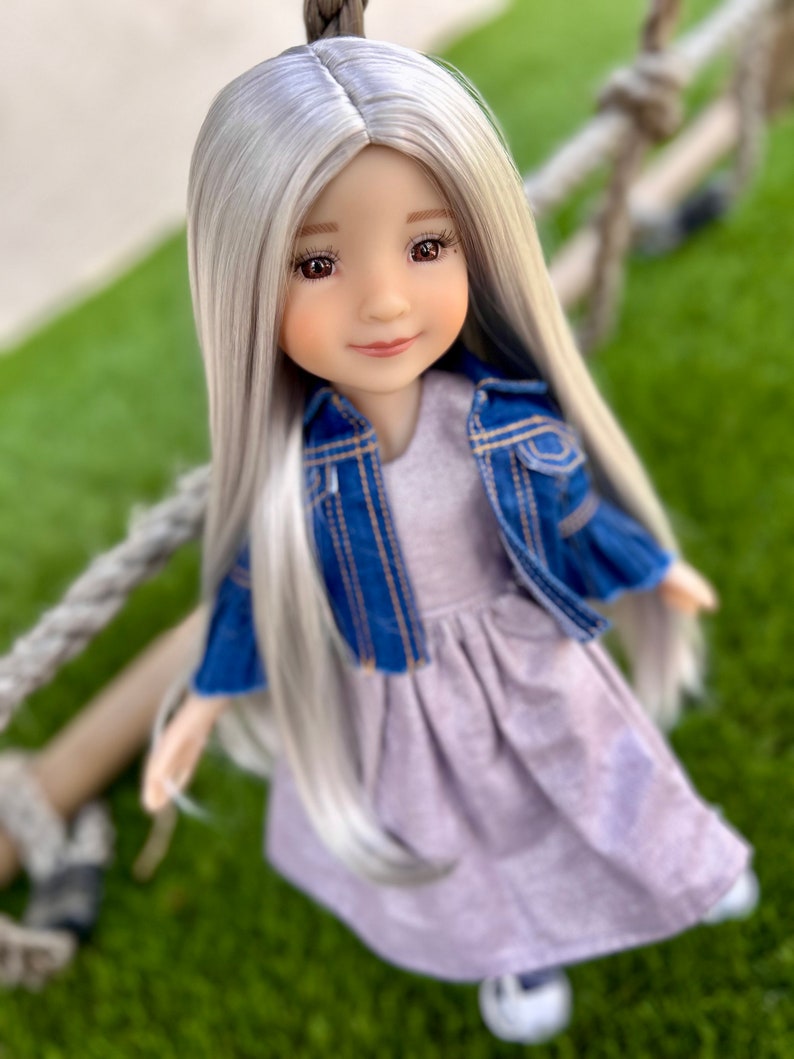 May include: A doll with long, silver hair and brown eyes, wearing a lavender dress and a blue denim jacket. The doll is posed outdoors on a green surface, with a rope in the background.