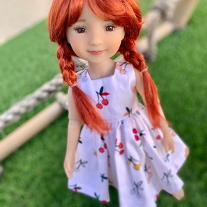 May include: A red-haired doll wearing a white dress with a cherry print. The doll has brown eyes and is looking directly at the camera.