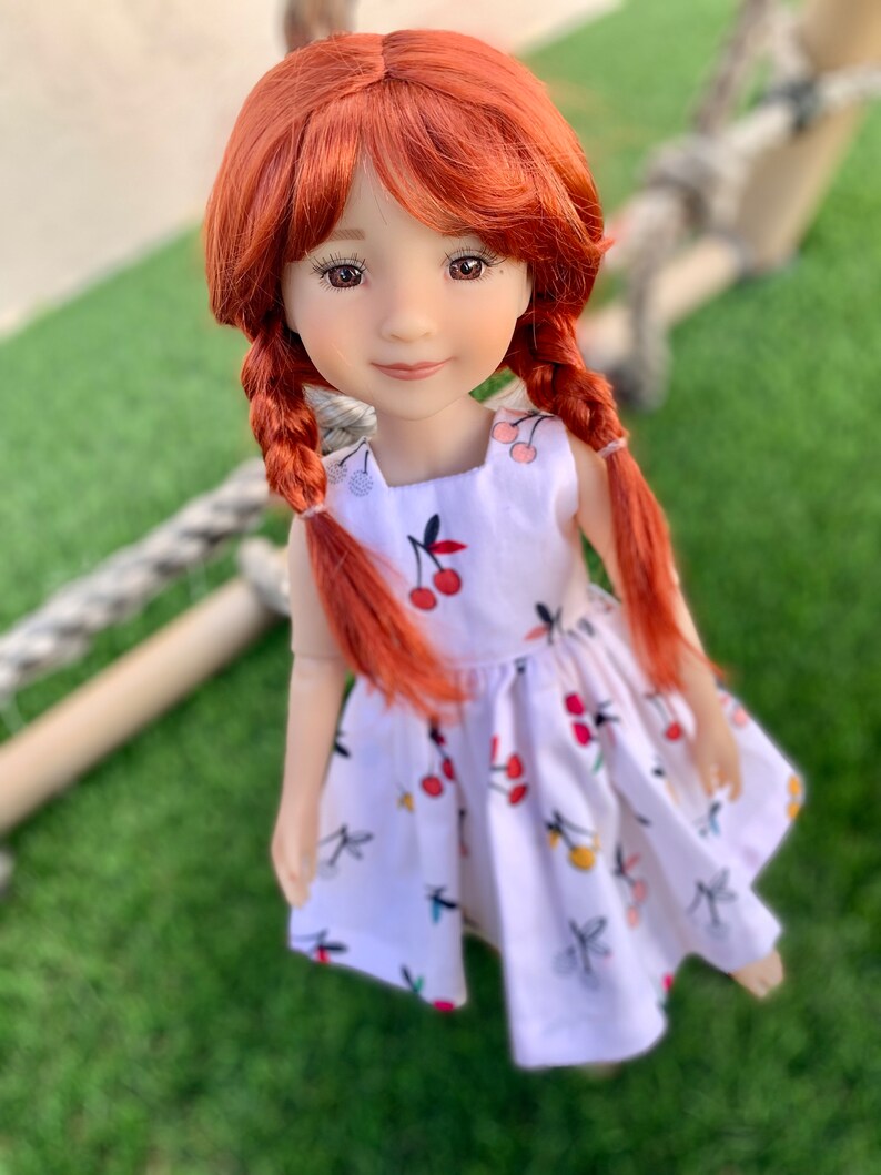 May include: A red-haired doll wearing a white dress with a cherry print. The doll has brown eyes and is looking directly at the camera.