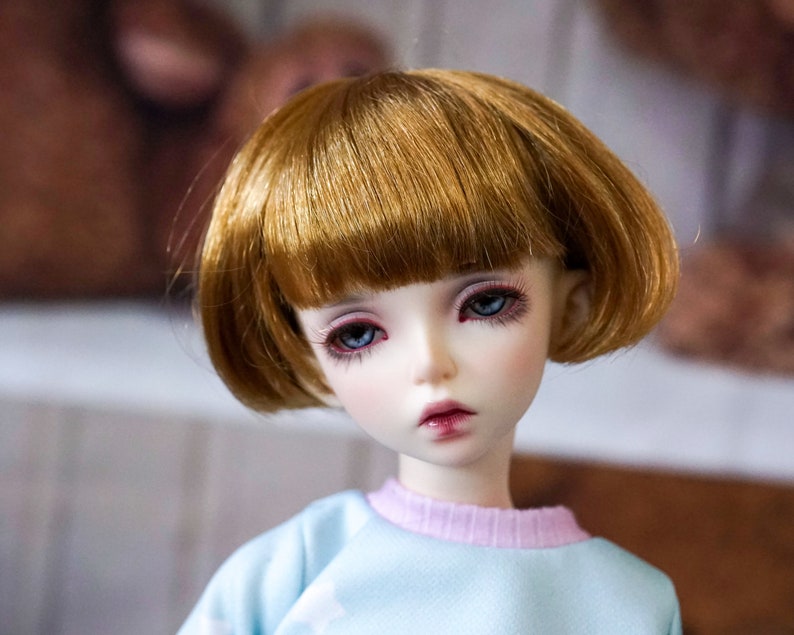 May include: A doll with short, brown hair and blue eyes. The doll is wearing a light blue shirt with a pink collar.