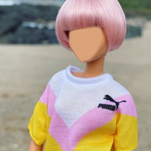 Peut inclure: Une poup&eacute;e portant un pull ray&eacute; jaune, rose et blanc avec un logo Puma noir. La poup&eacute;e a les cheveux roses coiff&eacute;s en bob.