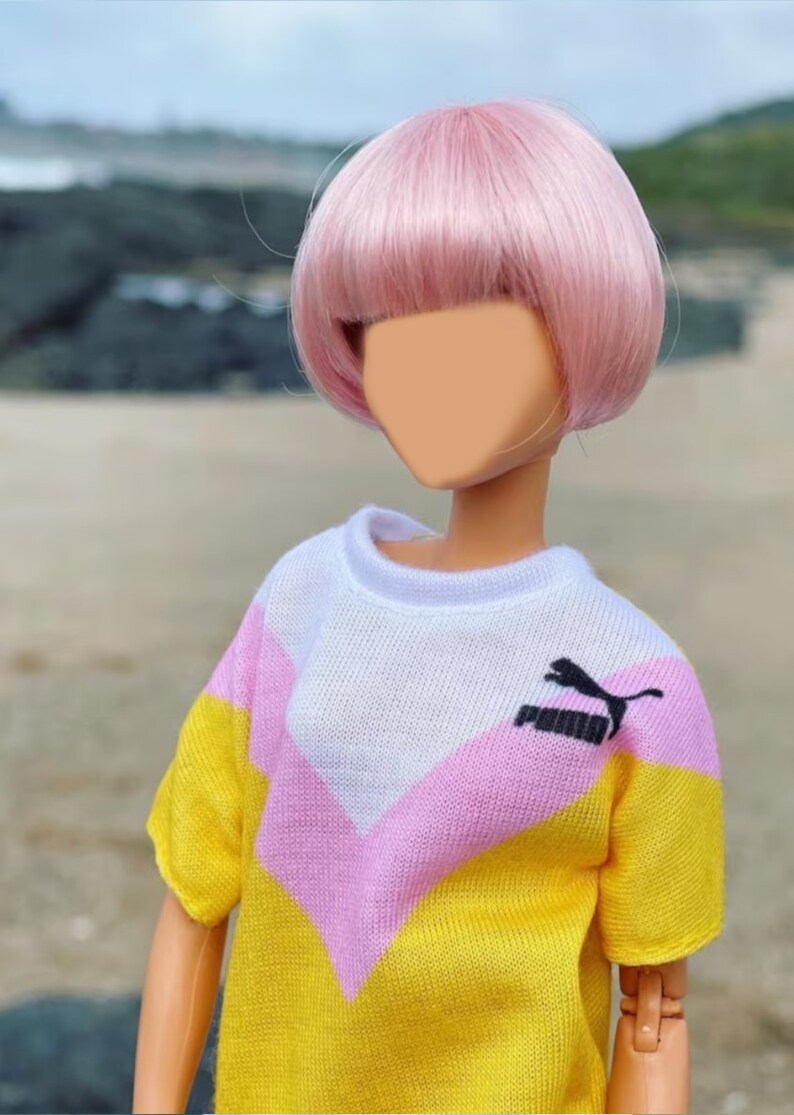 Peut inclure: Une poup&eacute;e portant un pull ray&eacute; jaune, rose et blanc avec un logo Puma noir. La poup&eacute;e a les cheveux roses coiff&eacute;s en bob.