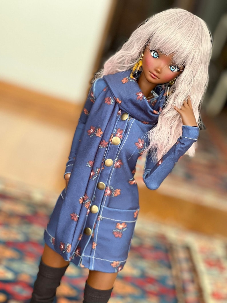 May include: A doll wearing a navy blue coat dress with a floral pattern in shades of orange and red. The dress features large gold buttons, pockets, and a matching scarf. The doll has long, light pink hair and is wearing black knee-high socks.