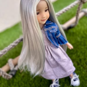 May include: A doll with long, silver hair, wearing a light purple dress and a blue denim jacket. The doll is seated on a rope swing, with white shoes and blue laces. The doll has a serene expression.