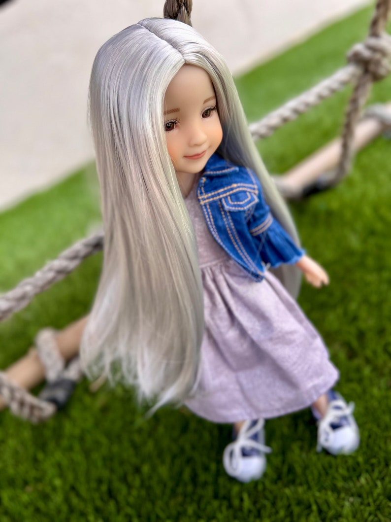 May include: A doll with long, silver hair, wearing a light purple dress and a blue denim jacket. The doll is seated on a rope swing, with white shoes and blue laces. The doll has a serene expression.