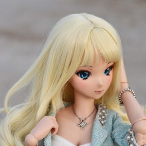 May include: A doll with long blonde hair, blue eyes, and a light blue jacket with pearl accents. She is wearing a white top and a silver necklace with a cross pendant.