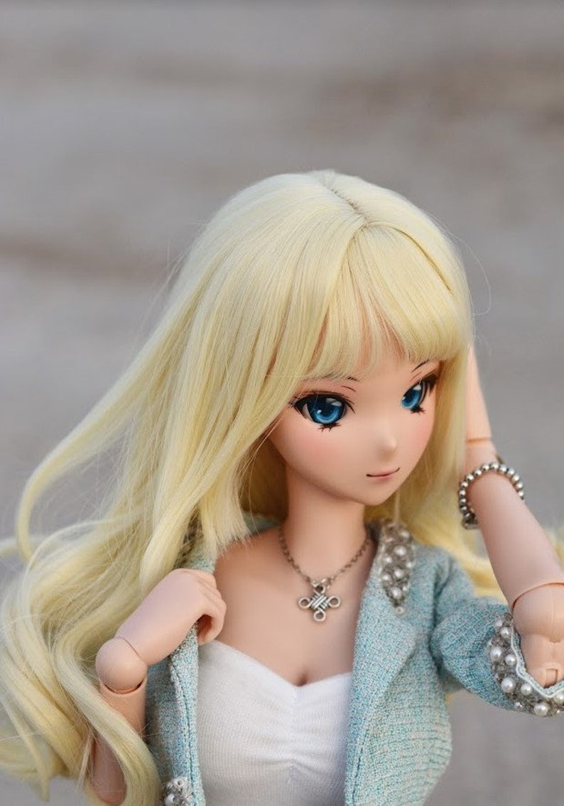 May include: A doll with long blonde hair, blue eyes, and a light blue jacket with pearl accents. She is wearing a white top and a silver necklace with a cross pendant.