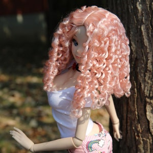 May include: A pink curly wig with a white top and colorful shorts. The doll has blue eyes and a pink blush.