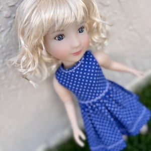 May include: A doll with shoulder-length blonde hair and blue eyes, wearing a sleeveless blue dress featuring a white polka dot pattern. The dress has a fitted bodice and a pleated skirt with a white stitched hem detail along the bottom edge.