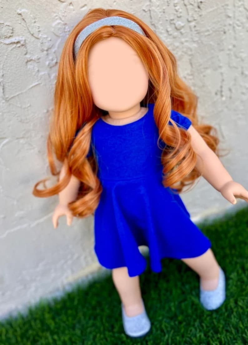May include: A doll with long, curly, auburn hair wearing a blue dress and a silver headband. The doll is standing on green grass.