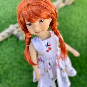 May include: A red-haired doll wearing a white dress with a cherry print. The doll has a sweet expression and is looking directly at the camera.
