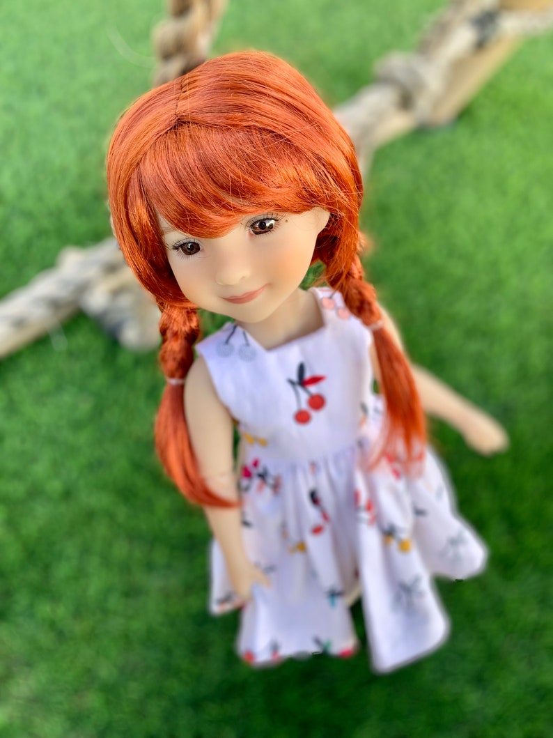 May include: A red-haired doll wearing a white dress with a cherry print. The doll has a sweet expression and is looking directly at the camera.