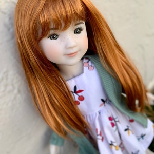 May include: A red-haired doll wearing a white dress with a cherry print and a sage green cardigan. The doll has freckles and blue eyes.