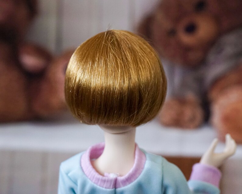May include: A doll with short, straight, light brown hair. The doll is wearing a light blue shirt with a pink collar.