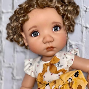 Puede incluir: Una muñeca con el pelo rizado marrón y ojos azules lleva un vestido blanco y amarillo con un lazo marrón. La muñeca está mirando a la cámara.