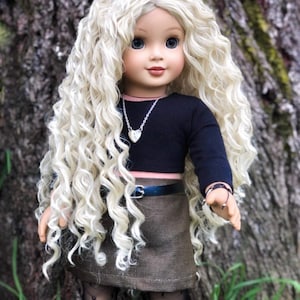 May include: A doll with long, curly blonde hair, blue eyes, and a fair complexion. The doll wears a black long-sleeved top, a brown skirt with a blue belt, and patterned tights. A silver necklace with a heart pendant and a ring are also visible.