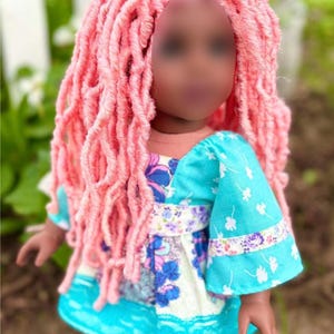 May include: A doll with long, textured pink hair styled in locs. The doll wears a short-sleeved dress featuring a bright turquoise fabric with a white floral pattern, accented by a band of purple and white floral print across the waist and sleeves.