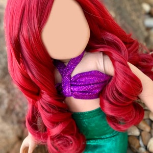 Puede incluir: Una peluca roja con el pelo largo y rizado, dise&ntilde;ada para una mu&ntilde;eca. La mu&ntilde;eca lleva un disfraz de sirena morado y verde.