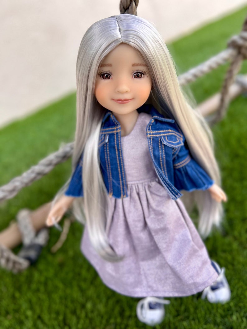 May include: A doll with long, silver hair and a denim jacket over a lavender dress. The doll is wearing white shoes and has a serene expression. The doll is sitting on a green surface.