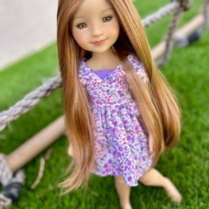 May include: A doll with long, light brown hair and a floral dress in shades of purple and white. The doll has a serene expression and is positioned on green grass, with a rope swing in the background.