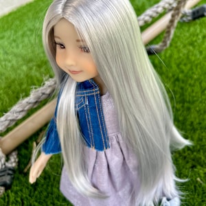 May include: A doll with long, silver hair, wearing a lavender dress and a blue denim jacket. The doll has a serene expression and is posed outdoors on a green surface, with a rope swing in the background.