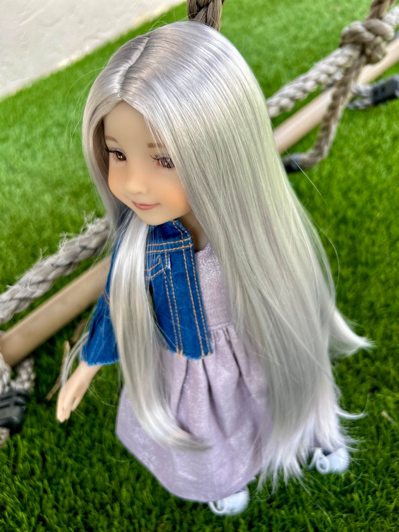 May include: A doll with long, silver hair, wearing a lavender dress and a blue denim jacket. The doll has a serene expression and is posed outdoors on a green surface, with a rope swing in the background.