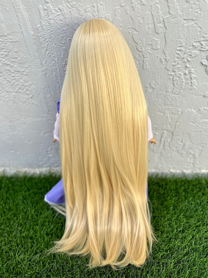 May include: A doll with long, flowing, blonde hair. The hair is styled straight and reaches the doll's waist. The doll is wearing a white top with a purple dress. The doll is kneeling on green grass in front of a textured gray wall.