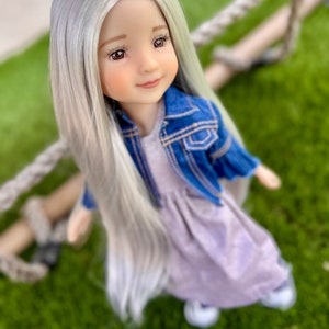 May include: A doll with long, silver-gray hair, wearing a light purple dress and a blue denim jacket. The doll has a serene expression and is posed against a backdrop of green grass and a rope swing.