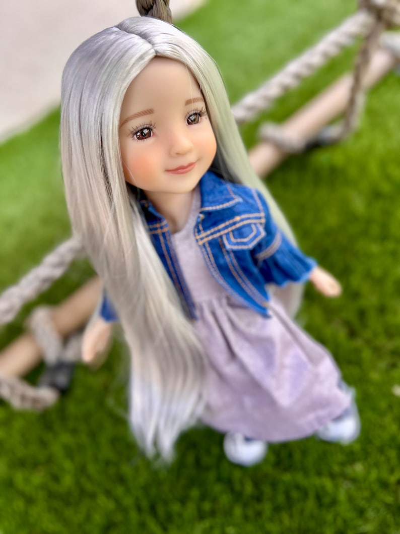 May include: A doll with long, silver-gray hair, wearing a light purple dress and a blue denim jacket. The doll has a serene expression and is posed against a backdrop of green grass and a rope swing.