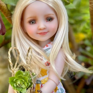 May include: A blonde doll with brown eyes wearing a yellow and white floral dress and a brown bracelet. The doll is holding a woven basket with a green succulent plant inside.